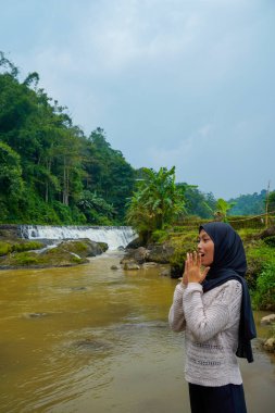 Asyalı Müslüman kadının, elleri önde, yüzü nehir kenarına dönük, şelale arkaplan ve reklam alanı için açık gökyüzü olan şok ifadesini görmek.