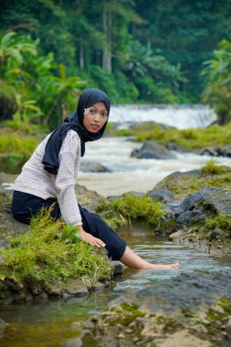 Tesettür takan Asyalı bir kadın, yeşil ağaçlar ve küçük bir şelaleyle ayaklarını suya sokarken kayalık bir nehrin kenarında oturur..