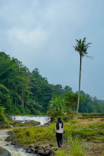 Dağ manzarası, küçük şelaleden akan nehirler ve önplanda duran kadın, tesetteki kayaların üzerinde akan kayaların yanında duran tesettür ve berrak gökyüzü.