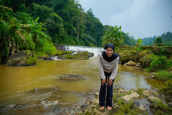 Arka planda şelaleyle nehir kıyısında eğilirken gülümseyen Asyalı Müslüman kadının ön görüntüsü.