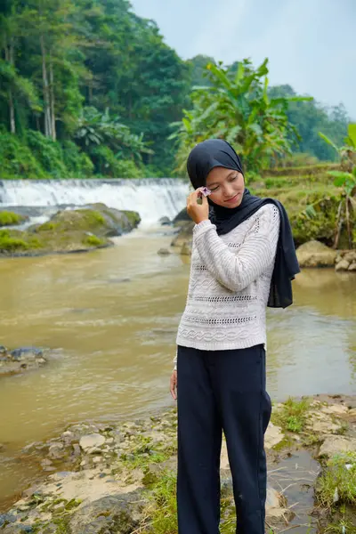 Nehir kıyısında tesettürlü Asyalı bir kadın elinde çiçekle bir şelalenin arka planına karşı duruyor..