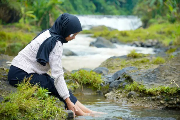Tesettürlü Asyalı bir kadın, yeşil ağaçlar ve küçük bir şelale ve reklam alanı için berrak bir gökyüzü ile suda yıkanmak için ayaklarını ıslatırken kayalık bir nehrin kenarında oturur..