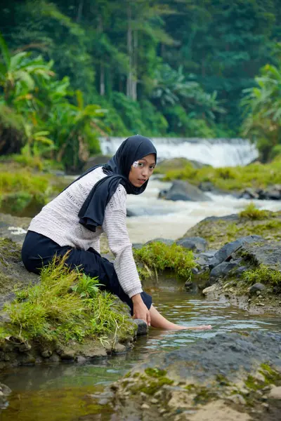 Tesettürlü Asyalı bir kadın, yeşil ağaçlar ve küçük bir şelale ve reklam alanı için berrak bir gökyüzü ile suda yıkanmak için ayaklarını ıslatırken kayalık bir nehrin kenarında oturur..