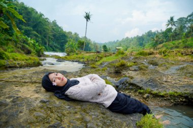 Asyalı Müslüman kadının yeşil ağaçlarla nehirdeki kayaların üzerinde yatışı ve reklam alanı için açık gökyüzü arkaplanı.