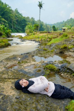 Asyalı Müslüman kadının yeşil ağaçlarla nehirdeki kayaların üzerinde yatışı ve reklam alanı için açık gökyüzü arkaplanı.