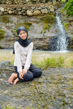 Asyalı Müslüman bir kadın kayalık bir nehir kıyısında oturuyor. Arkasında pirinç tarlaları ve küçük bir şelale var..