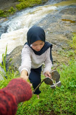 Tesettürlü bir kadın arka planda kayaların arasından akan bir nehirle tırmanmak için yardım istiyor..