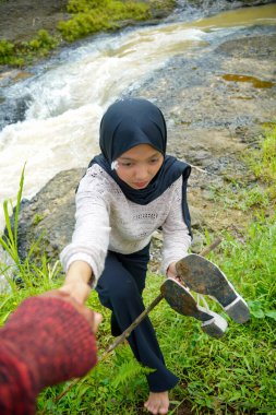 Tesettürlü bir kadın arka planda kayaların arasından akan bir nehirle tırmanmak için yardım istiyor..
