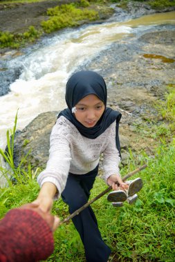 Tesettürlü bir kadın arka planda kayaların arasından akan bir nehirle tırmanmak için yardım istiyor..