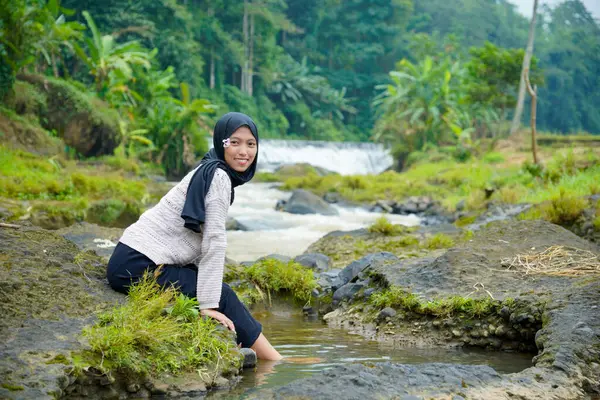 Tesettür takan Asyalı bir kadın, yeşil ağaçlar ve küçük bir şelaleyle ayaklarını suya sokarken kayalık bir nehrin kenarında oturur..