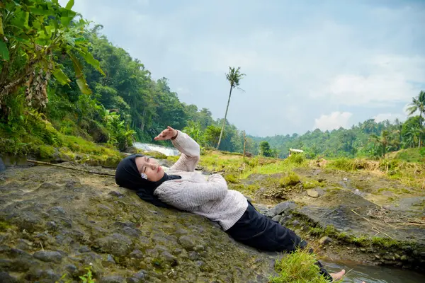 Asyalı Müslüman kadının yeşil ağaçlarla nehirdeki kayaların üzerinde yatışı ve reklam alanı için açık gökyüzü arkaplanı.