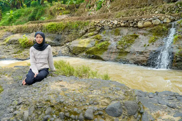 Asyalı Müslüman bir kadın kayalık bir nehir kıyısında oturuyor. Arkasında pirinç tarlaları ve küçük bir şelale var..