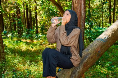 Asyalı Müslüman kadın ağaç gövdesinde oturmuş ormandaki bir şişeden su içiyor. Asyalı Müslüman kadın ormanda bir ağaç gövdesindeki şişede su içiyor..