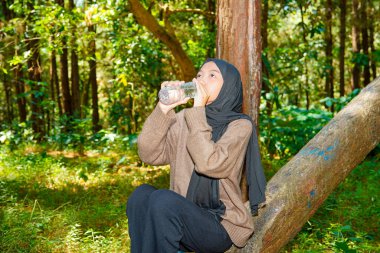Asyalı Müslüman kadın ağaç gövdesinde oturmuş ormandaki bir şişeden su içiyor. Asyalı Müslüman kadın ormanda bir ağaç gövdesindeki şişede su içiyor..