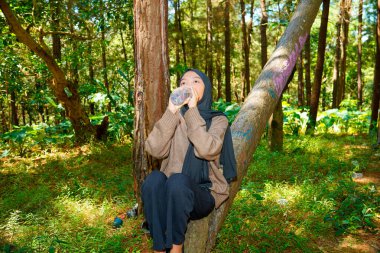 Asyalı Müslüman kadın ağaç gövdesinde oturmuş ormandaki bir şişeden su içiyor. Asyalı Müslüman kadın ormanda bir ağaç gövdesindeki şişede su içiyor..