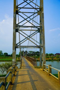 Reklam alanı için açık bir gökyüzü zeminine karşı demirden bir iskeleti olan yüksek bir asma köprü kulesi. Çelik bir asma köprü, parlak mavi gökyüzünün altında yeşilliklerle çevrili bir nehir boyunca uzanır..