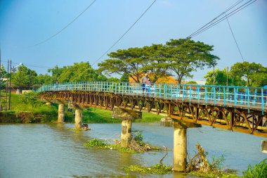 Jepara Central Java, 3 Haziran 2025, insanlar demir bir kiriş köprüsü üzerinde yürüyorlar mavi güvenlik korkulukları ile bir nehri geçiyor ağaçların arka planında ve temiz bir gökyüzü reklam alanı için.