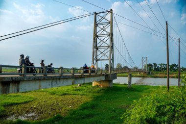 Jepara Central Java, 3 Haziran 2025, bir motosikletçi, reklam alanı için arka planda açık gökyüzü olan bir nehir üzerinden demir asma köprüsünü geçiyor..
