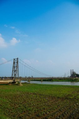 Nehir üzerindeki uzun asma köprü. Etrafı tarlalarla çevrili. Reklam alanı için temiz bir gökyüzü arkaplanı var..
