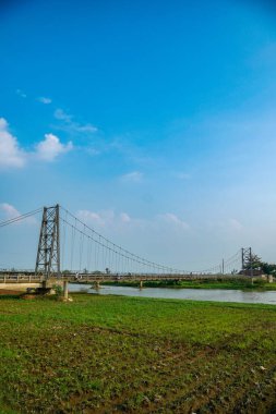 Nehir üzerindeki uzun asma köprü. Etrafı tarlalarla çevrili. Reklam alanı için temiz bir gökyüzü arkaplanı var..