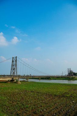 Nehir üzerindeki uzun asma köprü. Etrafı tarlalarla çevrili. Reklam alanı için temiz bir gökyüzü arkaplanı var..