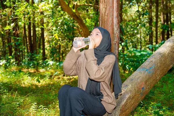 Asyalı Müslüman kadın ağaç gövdesinde oturmuş ormandaki bir şişeden su içiyor. Asyalı Müslüman kadın ormanda bir ağaç gövdesindeki şişede su içiyor..