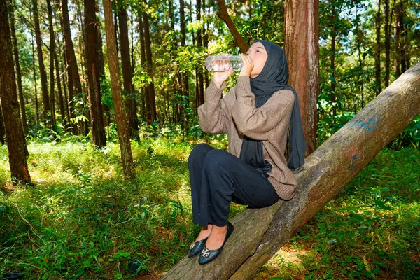 Bir kadının tesettürde dikiz aynasından bir çam ağacının yanına çömelmiş, işaret parmağı soyulmuş ağaç gövdesine dokunarak kauçuk özsuyu topluyor.