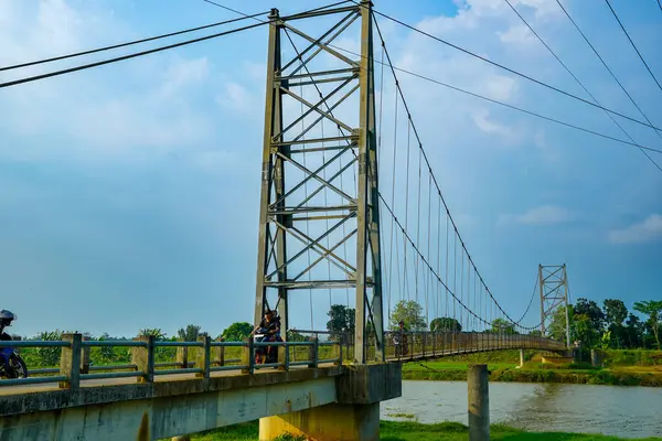Jepara Central Java, 3 Haziran 2025, bir motosikletçi, reklam alanı için arka planda açık gökyüzü olan bir nehir üzerinden demir asma köprüsünü geçiyor..