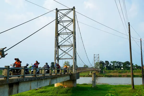 Jepara Central Java, 3 Haziran 2025, bir motosikletçi, reklam alanı için arka planda açık gökyüzü olan bir nehir üzerinden demir asma köprüsünü geçiyor..