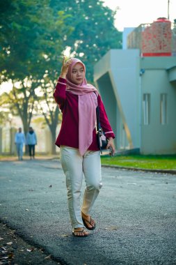 Asyalı Müslüman kadın hafifçe gülümseyerek binanın arka bahçesinde yürüyor, güzel Asyalı Müslüman kadın gülümsüyor ve kameraya bakıyor, güzel Asyalı genç Malezyalı kadın tesettürlü