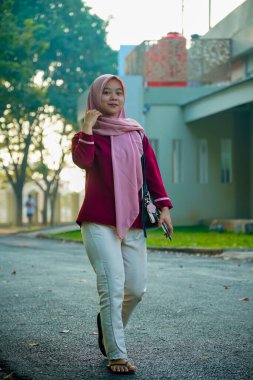 Asyalı Müslüman kadın hafifçe gülümseyerek binanın arka bahçesinde yürüyor, güzel Asyalı Müslüman kadın gülümsüyor ve kameraya bakıyor, güzel Asyalı genç Malezyalı kadın tesettürlü