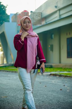 Asyalı Müslüman kadın hafifçe gülümseyerek binanın arka bahçesinde yürüyor, güzel Asyalı Müslüman kadın gülümsüyor ve kameraya bakıyor, güzel Asyalı genç Malezyalı kadın tesettürlü