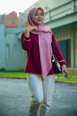 Asyalı Müslüman kadın hafifçe gülümseyerek binanın arka bahçesinde yürüyor, güzel Asyalı Müslüman kadın gülümsüyor ve kameraya bakıyor, güzel Asyalı genç Malezyalı kadın tesettürlü