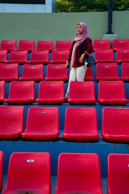 Asyalı Müslüman kadın bir futbol stadyumunda kırmızı seyirci koltukları arasında duruyor.