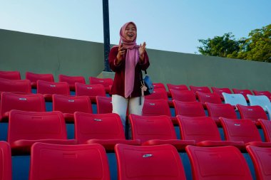 Asyalı Müslüman kadın bir futbol stadyumunda kırmızı seyirci koltuklarının arasında duruyor. Boş kırmızı stadyum koltukları arasında neşeli bir kadın destek, spor anlayışı ve sağlıklı yaşam tarzı için gülümsüyor..