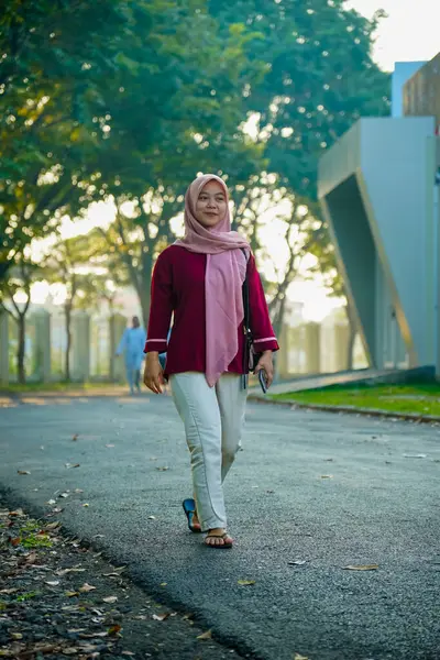 Asyalı Müslüman kadın hafifçe gülümseyerek binanın arka bahçesinde yürüyor, güzel Asyalı Müslüman kadın gülümsüyor ve kameraya bakıyor, güzel Asyalı genç Malezyalı kadın tesettürlü