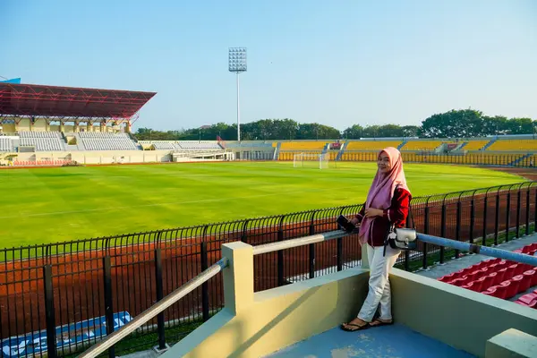 Stadyumdaki sahada duran Asyalı Müslüman kadın, mavi gökyüzü ve bulutlu Asya futbol stadyumu, stadyum arka planında küçük çantalı kırmızı elbiseli güzel kız, ayakta duran Müslüman kadın.
