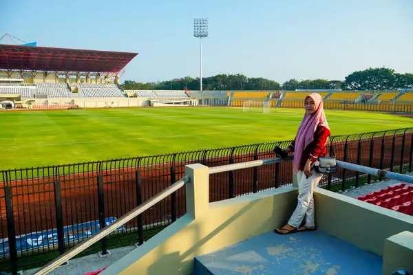 Stadyumdaki sahada duran Asyalı Müslüman kadın, mavi gökyüzü ve bulutlu Asya futbol stadyumu, stadyum arka planında küçük çantalı kırmızı elbiseli güzel kız, ayakta duran Müslüman kadın.