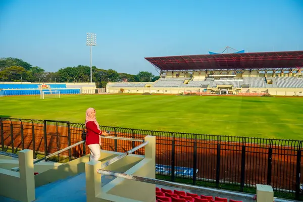 Stadyumdaki sahada duran Asyalı Müslüman kadın, mavi gökyüzü ve bulutlu Asya futbol stadyumu, stadyum arka planında küçük çantalı kırmızı elbiseli güzel kız, ayakta duran Müslüman kadın.