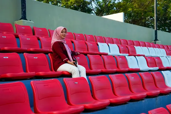 Asyalı Müslüman kadın kırmızı stadyum koltuklarında oturuyor, pembe tesettür ve kestane rengi elbise giyen kadın rahat bir ifadeyle kırmızı stadyum koltuğunda oturuyor..