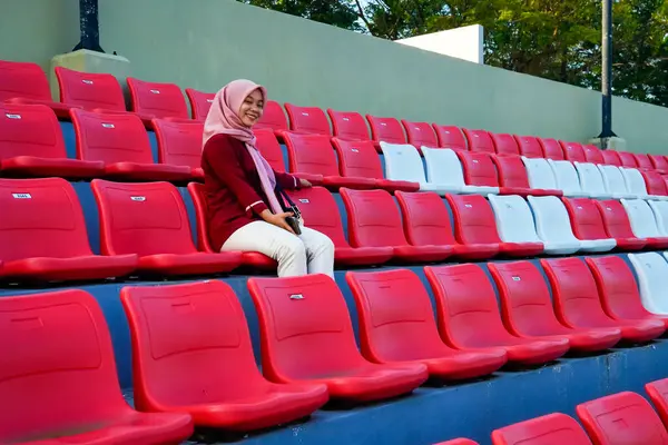 Asyalı Müslüman kadın kırmızı stadyum koltuklarında oturuyor, pembe tesettür ve kestane rengi elbise giyen kadın kameraya gülümseyen bir ifadeyle kırmızı stadyum koltuğunda oturuyor..