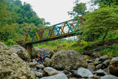Jepara Central Java, 9 Haziran 2025, köprünün altında bir grup çocuğun oynadığı kayalık bir nehrin üzerinde renkli bir çelik köprü uzanıyor.