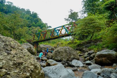 Jepara Central Java, 9 Haziran 2025, köprünün altında bir grup çocuğun oynadığı kayalık bir nehrin üzerinde renkli bir çelik köprü uzanıyor.