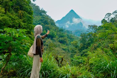 tesettürlü bir kadının yan görüntüsü dağlık bir manzaranın ortasındaki sis kaplı bir dağın zirvesini gösteriyor.