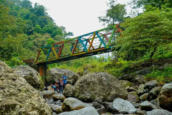 Jepara Central Java, 9 Haziran 2025, köprünün altında bir grup çocuğun oynadığı kayalık bir nehrin üzerinde renkli bir çelik köprü uzanıyor.