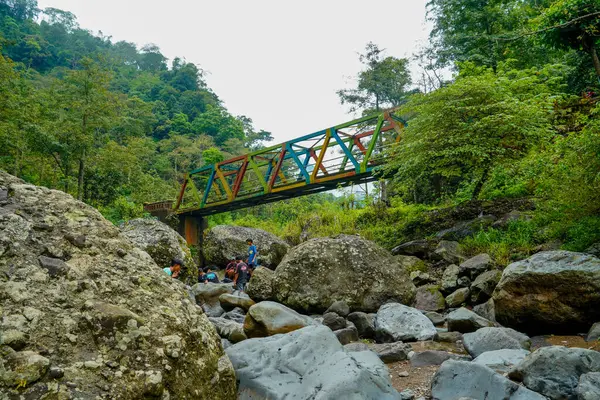 Jepara Central Java, 9 Haziran 2025, köprünün altında bir grup çocuğun oynadığı kayalık bir nehrin üzerinde renkli bir çelik köprü uzanıyor.