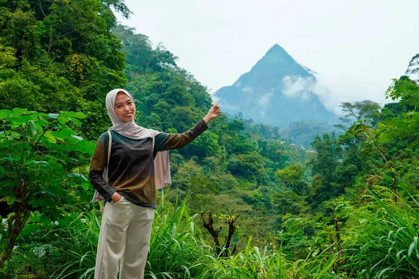 Asyalı Müslüman kadın, dağlık bir arazinin ortasındaki sis kaplı bir dağ zirvesini işaret ediyor.