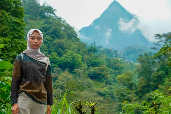 Asyalı Müslüman kadın kameraya bakıyor. Arka planda sisli dağ zirveleri var. Dağlık arazinin ortasında.