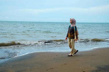 Kumsalda yürüyen tesettürlü Cava 'lı kadınlar, sahilde yürüyen Asyalı Müslüman kadınlar reklam alanı için küçük dalgalar ve boş gökyüzü ile çarpışıyorlar..