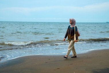 Kumsalda yürüyen tesettürlü Cava 'lı kadınlar, sahilde yürüyen Asyalı Müslüman kadınlar reklam alanı için küçük dalgalar ve boş gökyüzü ile çarpışıyorlar..